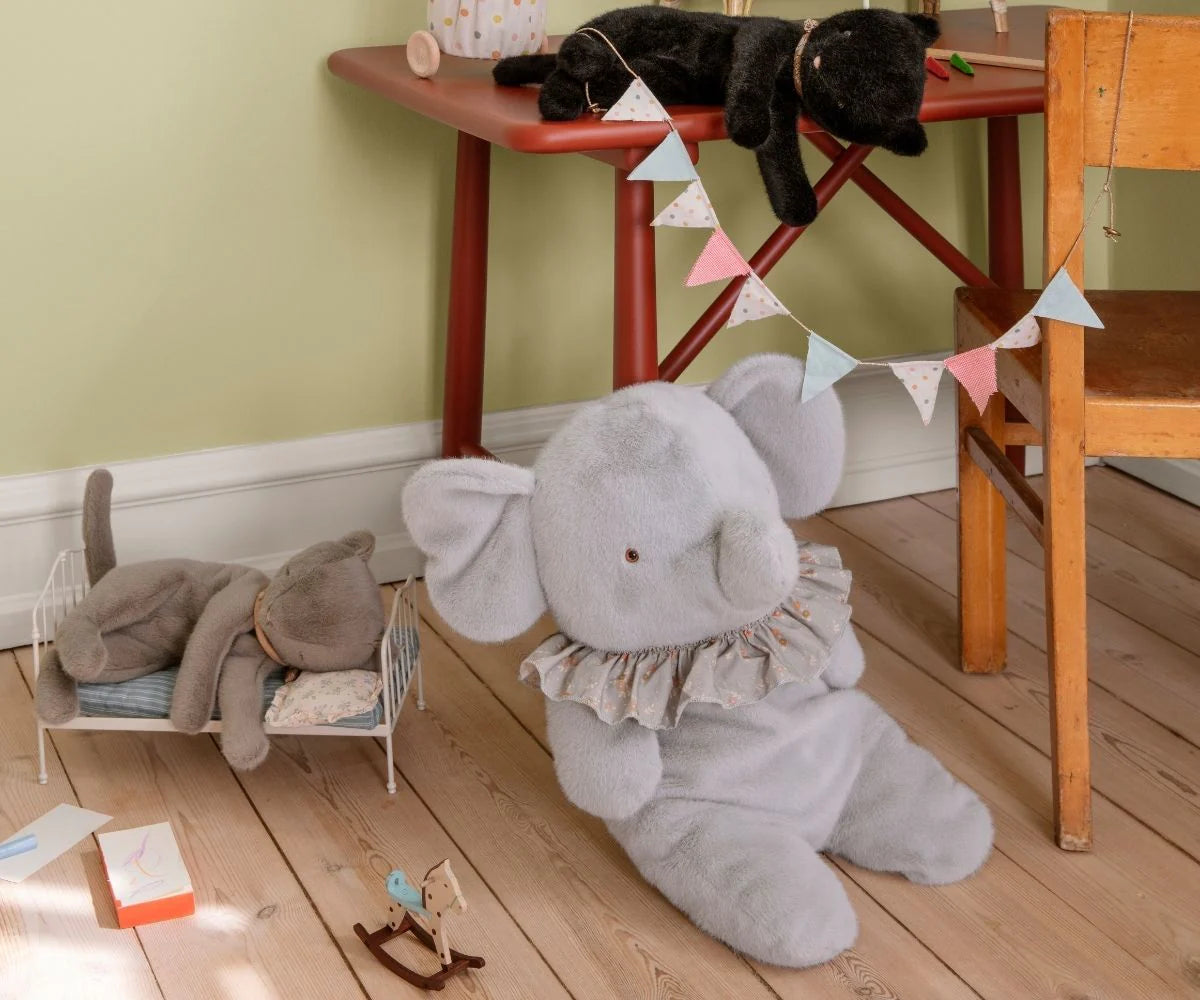 Children's room with a large gray elephant plush toy on the floor, a small dollhouse, and a black cat on a red table. | Maileg UK