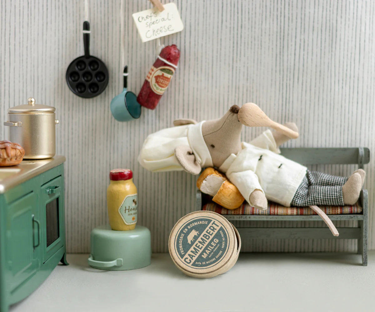 Toy chef mouse lying on a small bed in a miniature kitchen setting with various kitchen items around. |Chef clothes, Big sister and big brother mouse | Maileg UK
