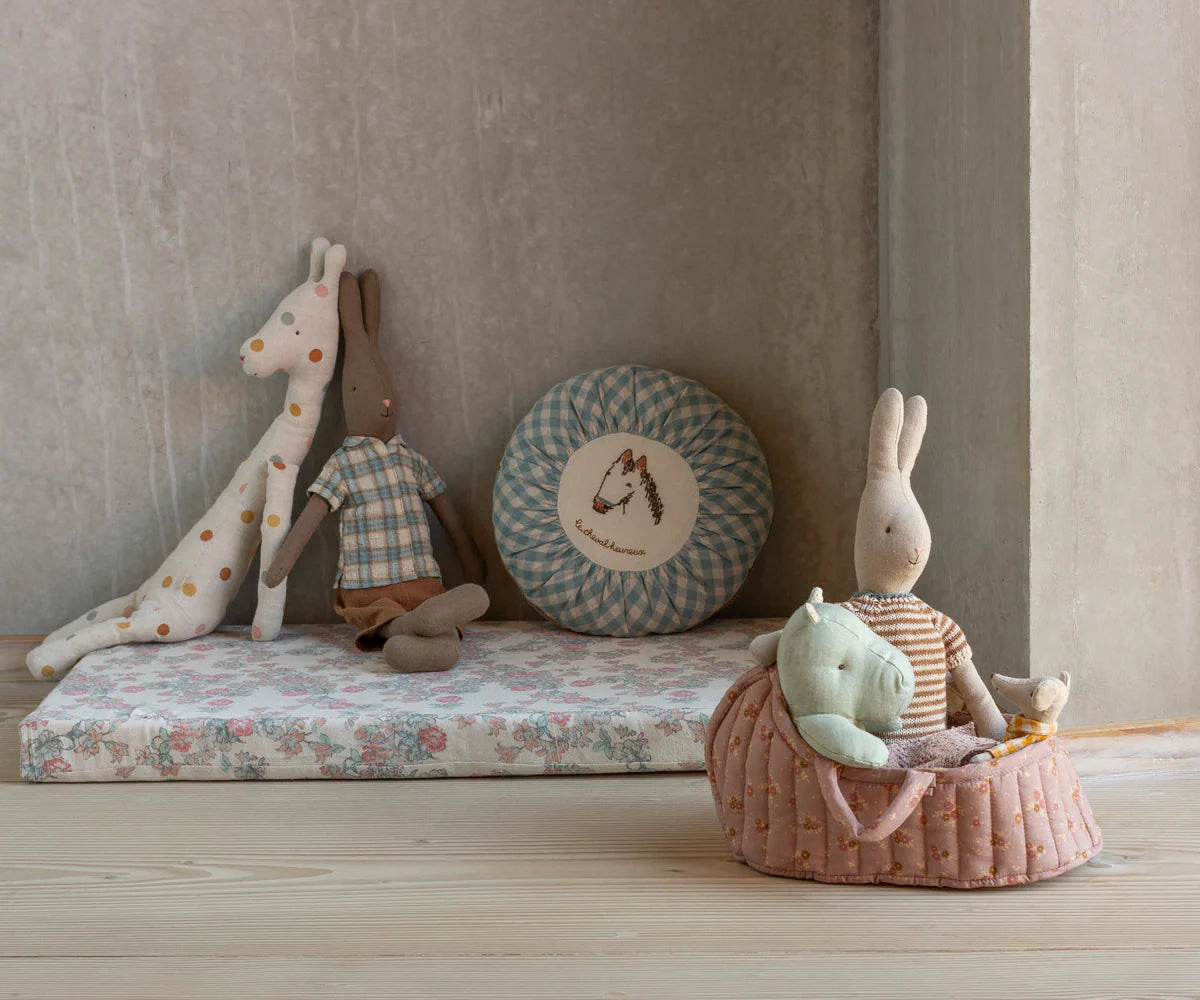 Children's toys including a giraffe, rabbit, and other plush items on a floral mattress against a neutral wall. | Maileg UK