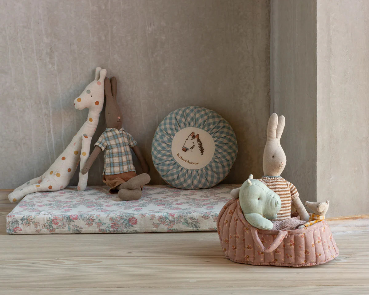 Children's toys including a giraffe, rabbit, and other plush items on a floral mattress against a neutral wall. | Maileg UK