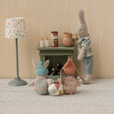 Toy figures of a rabbit, bear, and gnome around a small table with a lamp and jars on a textured surface.