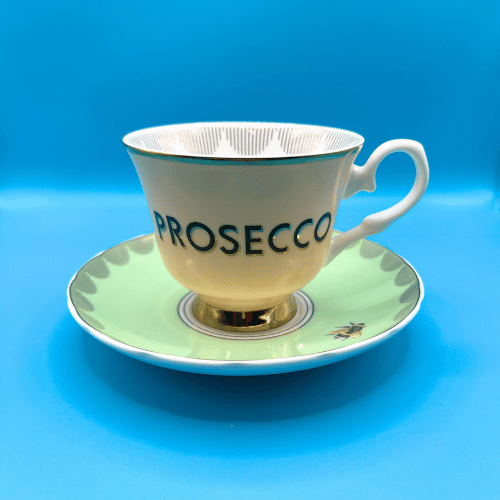 yvonne ellen bone chine cup ad saucer with gold detailing. A peach china cup with the cheeky slogan prosecco sits on a dainty green saucer with an art deco edging and bumble bee 