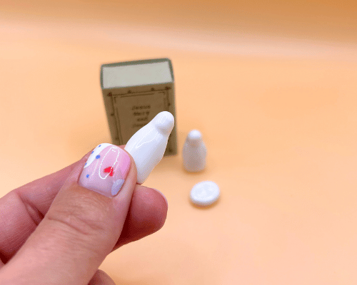 Small figurines and a miniature matchbox on a peach-colored surface with a hand holding a white figurine.