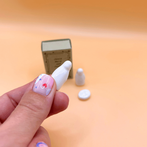 Small figurines and a miniature matchbox on a peach-colored surface with a hand holding a white figurine.